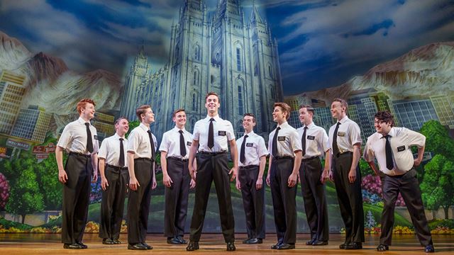 The cast of The Book of Mormon smiling together in front of a background of an old cathedral.