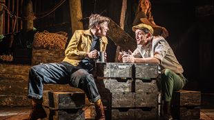 On the stage of The Curious Case of Benjamin Button, two men sit on crates and drink at a table made of wooden boxes.