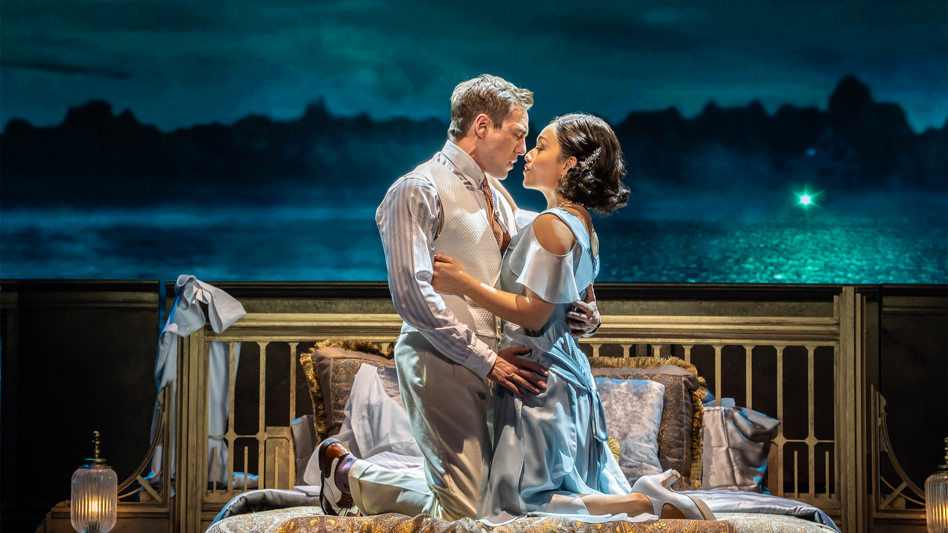 Jamie Muscato and Frances Mayli McCann in The Great Gatsby at the London Coliseum. Image courtesy of London Theatre Direct/Johan Persson. Two actors embrace on top of a bed against a coastal backdrop.