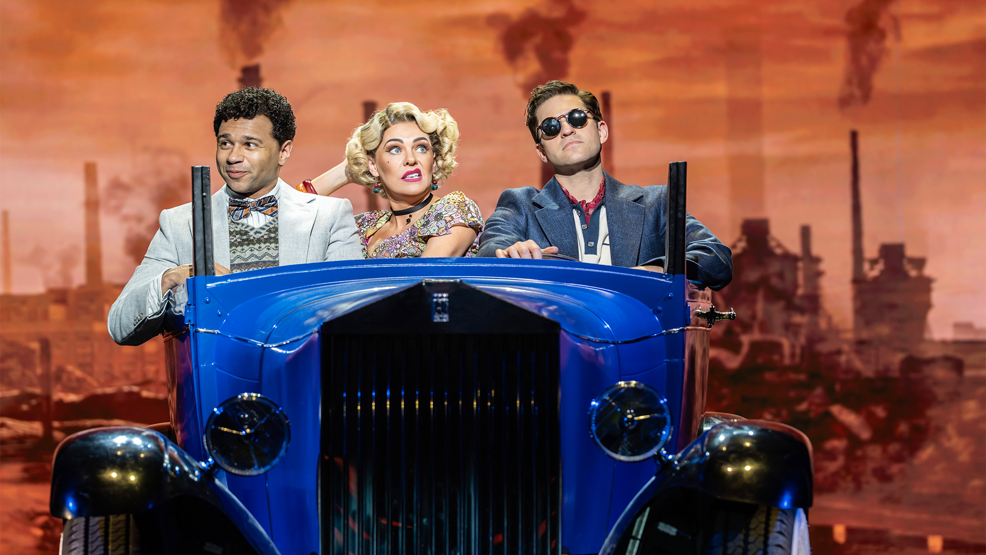 Corbin Bleu, Rachel Tucker and Jon Robyns in The Great Gatsby at the London Coliseum. Image courtesy of London Theatre Direct/Johan Persson. Three actors pictured in an old school car against a city backdrop in a West End show
