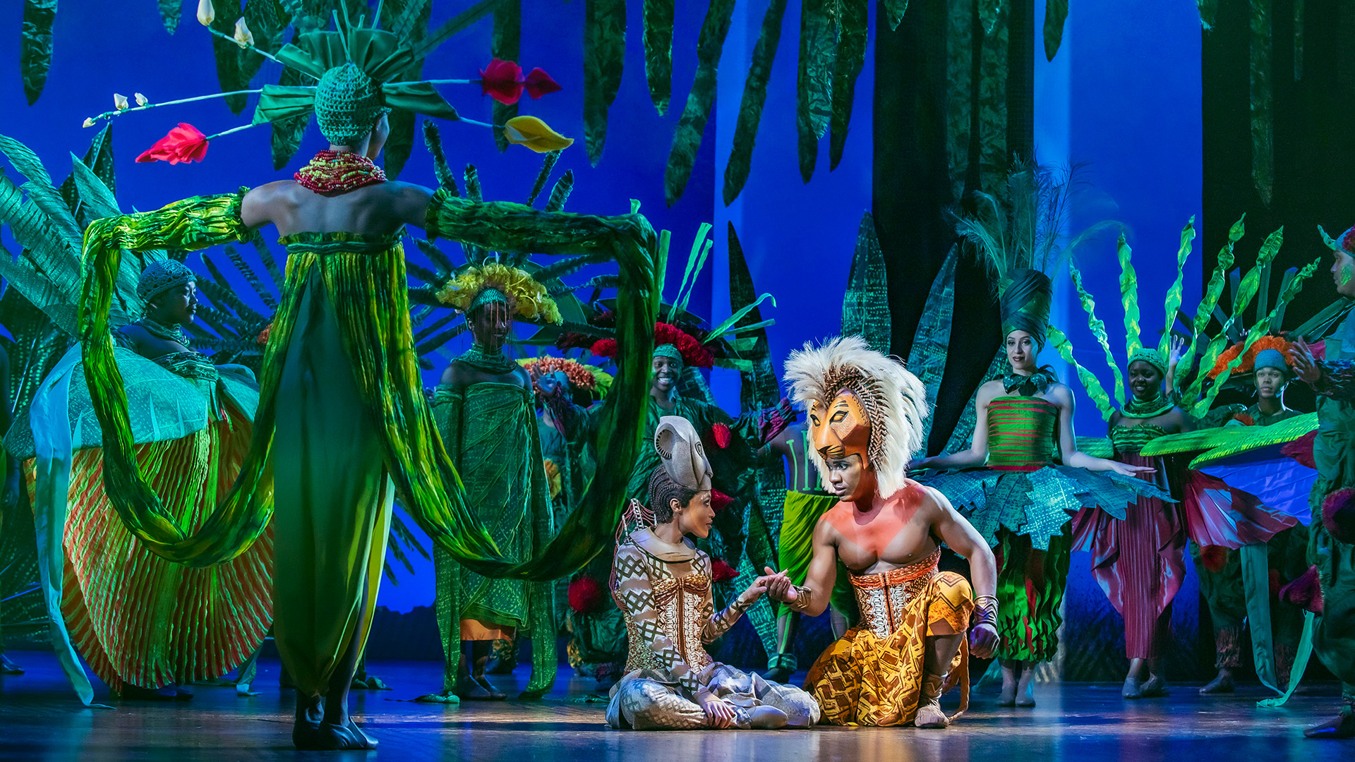 Revisit one of Disney's best-loved animated films, The Lion King, with its long-running musical theatre production at the Lyceum Theatre. Image courtesy of Disney. Actors portraying Simba and Nala hold hands while other members of The Lion King musical cast surround them, dressed and posing as luxurious and tropical trees and plants.