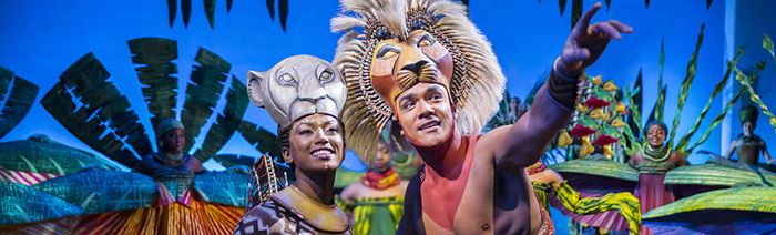 The actor playing Simba in The Lion King with a lion's head is talking to Lana and showing her something on the horizon. They are surrounded by the cast in colourful costumes in the background.