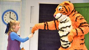 A life size tiger faces a delighted young girl wearing a purple dress carrying a plush toy.