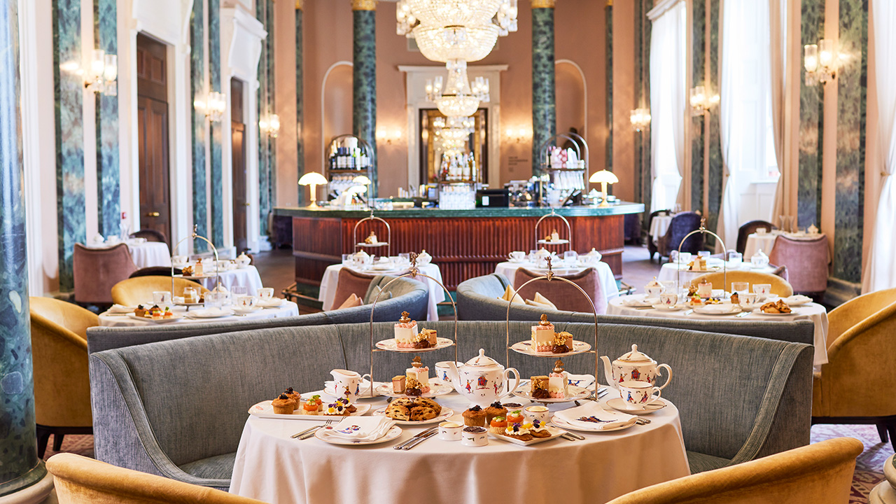 A table filled with delicious sweet treats and glasses of champagne and the Theatre Royal Drury Lane Champagne Afternoon tea in the Grand Saloon. A grand room with exquisite blue and yellow furnishings with a large white table laid out with a traditional afternoon tea setting.