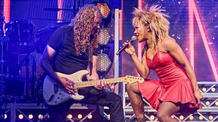 west end theatre stage with red and blue atmospheric lights; a man playing the guitar and a woman crouched over singing alongside him