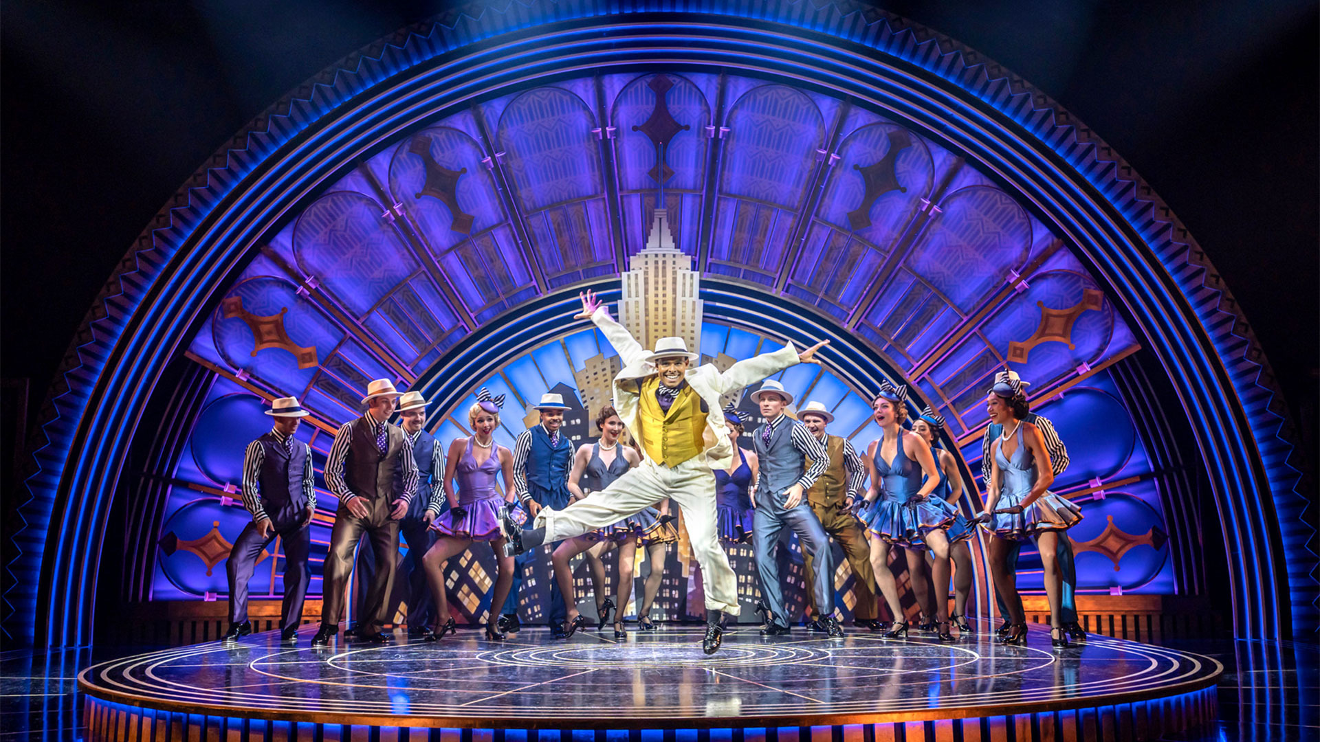 Dive into the golden years of Hollywood with the return of the musical sensation Top Hat, showing at the Southbank Centre. Image courtesy of Storyhouse PR/ photo credit: Johan Persson. Dressed in a three-piece suit, Phillip Attmore is jumping and dancing on stage with the rest of the Top Hat cast, who are standing at the back.