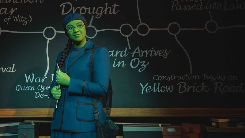 Elphaba in Wicked the musical at the Apollo Victoria Theatre. Photo credit: London Theatre Direct/ Matt Crockett. Elphaba wears the school uniform standing in front of a black board.