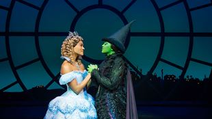 The actress portraying Glinda in Wicked the musical is seen wearing blue ball gown while holding hands witht eh actress playing the role of Alphaba, famous for her green skin and wearing a witch hat and all black outfit.
