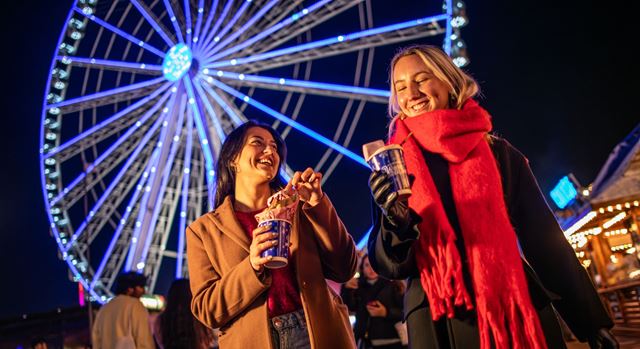Two people stand in coats in front of a ferris wheel eating hot chocolate.