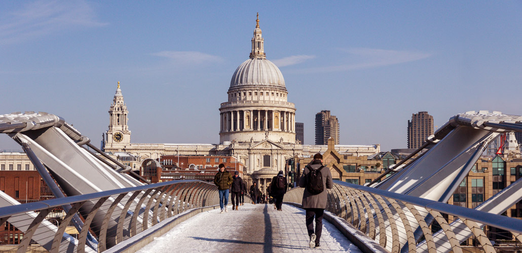 People walking across the London Millennium Bridge in the snow against the backdrop of St Paul's Cathedral.