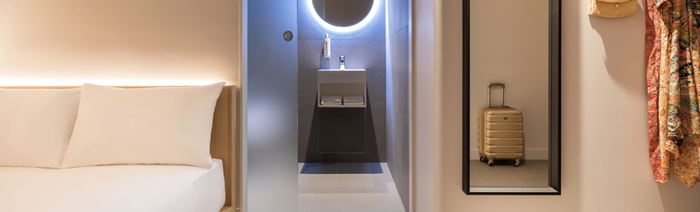 A warm and modern room with wooden-framed double bed, long black mirror and open plan walkway to the bathroom with a light up round mirror