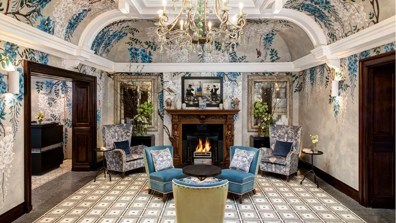 Be greeted with opulance the moment you walk through the front doors of Brown's. Photo: Janos Grapow. Image courtesy of Rocco Forte Hotels. A sumptuous hall decorated in blue and silver with velvet chairs in front of a fire and a chandelier glittering above the chairs.