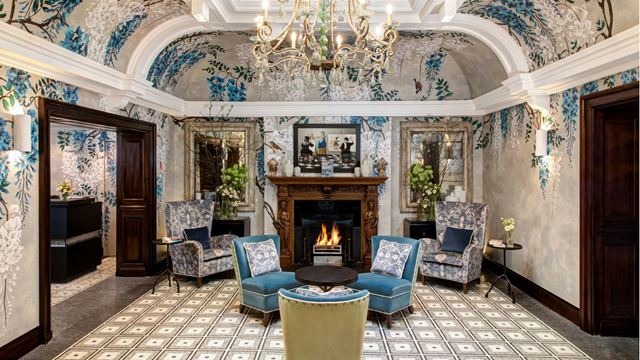 A sumptuous hall decorated in blue and silver with velvet chairs in front of a fire and a chandelier glittering above the chairs.