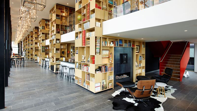 A photo of a communal area at Citizen M Tower of London with large wooden bookshelves with an area in the foreground featuring an Eames lounge chair, table and rug with stairs going up to the next level