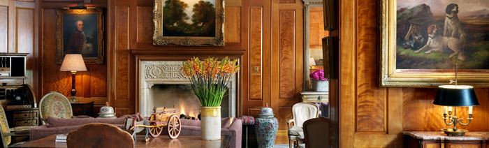 Gold framed classical paintings hang on dark wooden walls in the drawing room with a crackling fireplace, armchairs and table