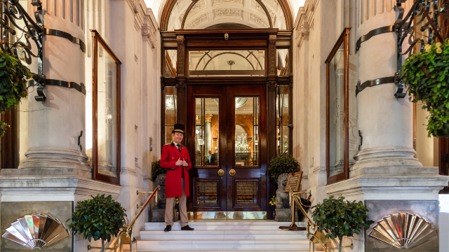 A smiling doorman dressed in an elegant red uniform is standing by the grand entrance of the Mandarin Oriental Hyde Park in Knightsbridge.