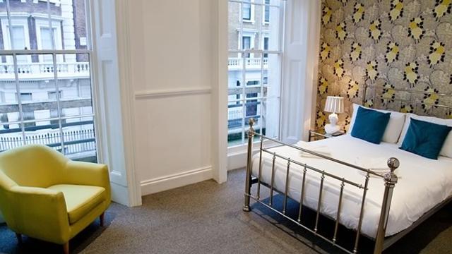 Room at Georgian House Hotel. Image courtesy of Georgian House Hotel. A double bed with white linen and blue cushions, with statement wallpaper behind and a pale green chair in the corner facing the bed.