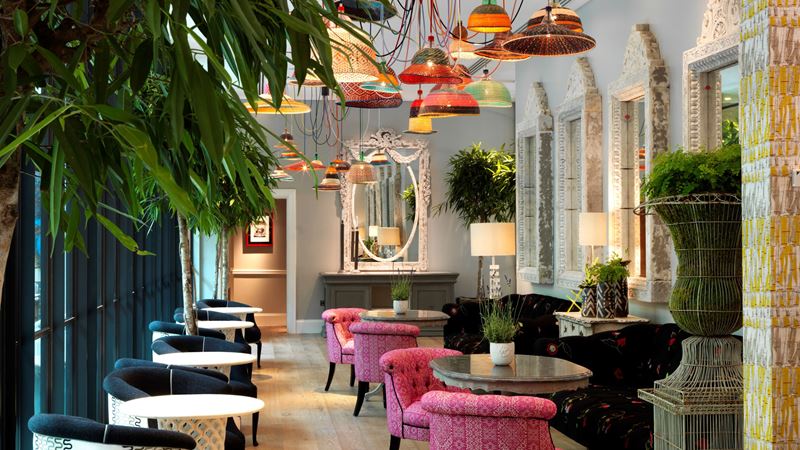 Unwind in the luxurious Orangery at Ham Yard Hotel. Credit: Firmdale Hotels. Image courtesy of Firmdale Hotels. Colourful pendant lamps hang above patterned chairs and tables with plants lining the room