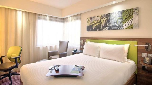 Double room at Hampton by Hilton London Croydon. Credit: Hampton by Hilton London Croydon. Image courtesy of Hampton by Hilton London Croydon. A bed with a lime green headboard, London-themed picture and a yellow chair.