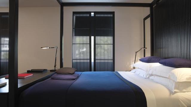 Modern room at La Suite West. Credit: La Suite West. Image courtesy of La Suite West. Looking across a double bed towards windows. The bed has a blue duvet and white linen, with a black headboard matching the blinds on the window.