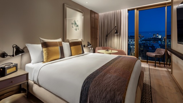 The Londoner Hotel. Image courtesy of Edwardian Hotels London. A room at The Londoner Hotel, with a large bed and views over London at night from a floor-to-ceiling window.