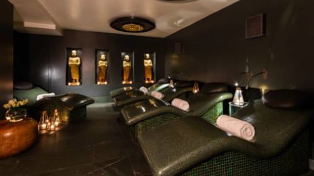 The dark, stylish May Fair Spa at May Fair Hotel, featuring dark green seating, dark-coloured wall and illuminated gold-coloured sculptures.