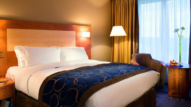 Sofitel London Gatwick North. Image courtesy of Sofitel A room with a large bed, navy blue throw and standard lamp, with net curtains drawn and a window behind.