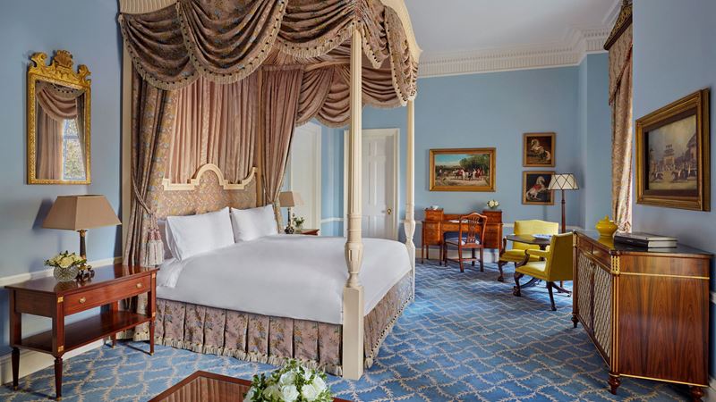 Feel like royalty in the Presidential Suite at The Lanesborough. Credit: The Lanesborough. Image courtesy of Perowne International. a four poster bed with golden gathered framing stands in the middle of a room lined with dark wooden furniture and gold framed paintings