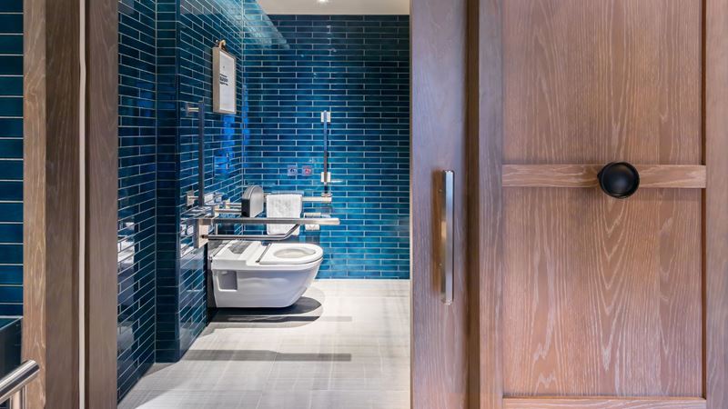 Relax in state-of-the-art accessible rooms at The Londoner with features such as handrails and level access to ensure a comfortable stay. Credit: The Londoner. Image courtesy of The Londoner. A view of the private bathroom inside an accessible room at The Londoner with modern blue tiles, wooden doors and accessible handrails around the toilet