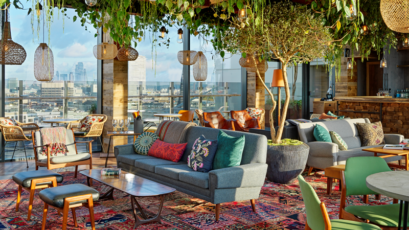 Trailing plants hang from the ceiling of the Nest lounge at Treehouse London with red Persian rugs, wooden hanging pendant lights and comfy chairs and sofas with floor-to-ceiling windows boasting a view of London's cityscape